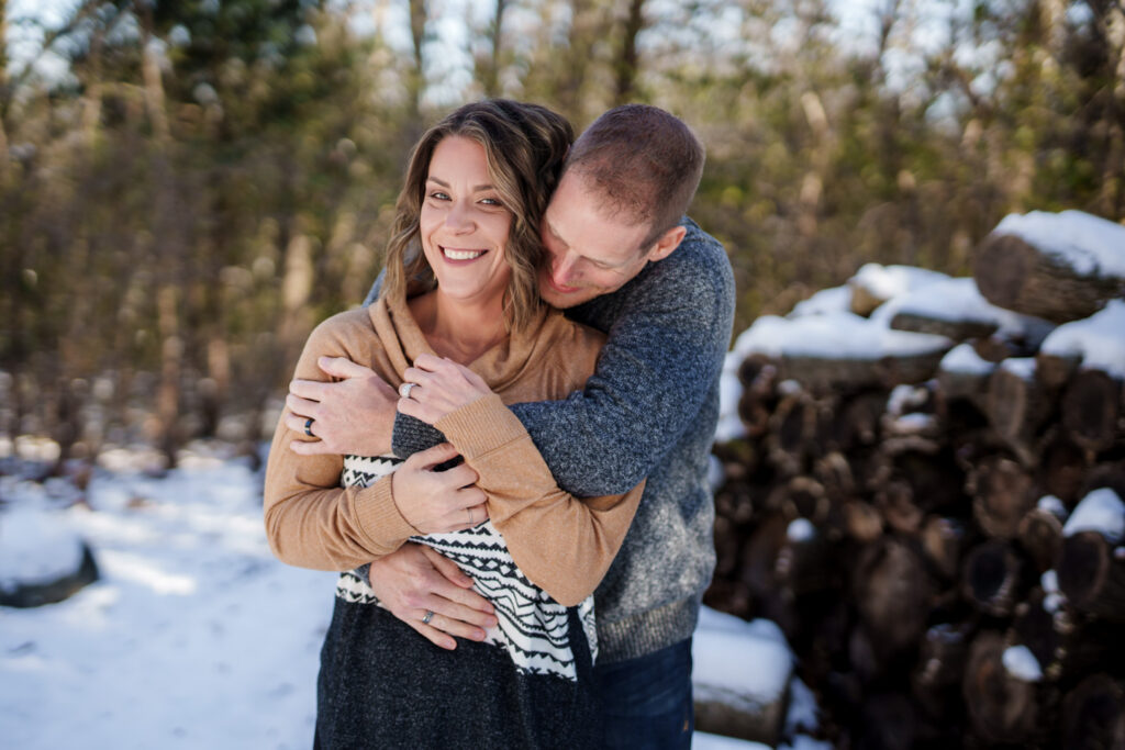 Jen and Mark Heller hugging in front of a wood pile 